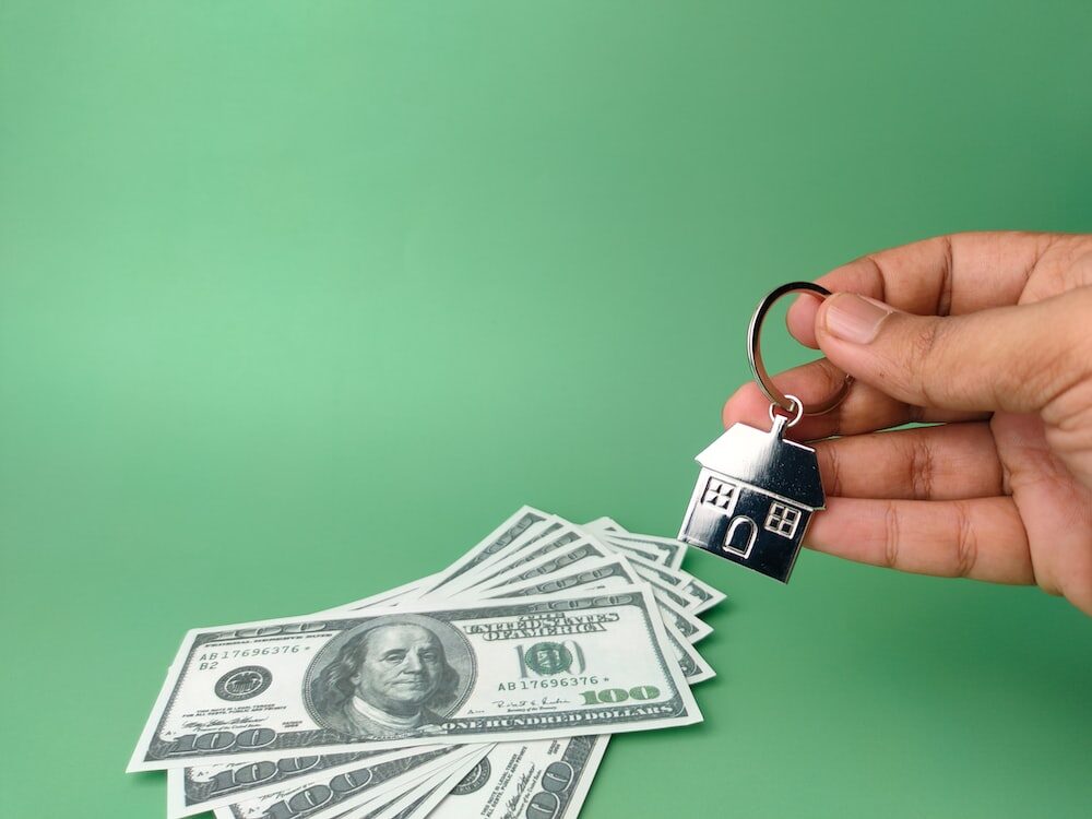 A person holding a house-shaped keychain above a stack of hundred-dollar bills