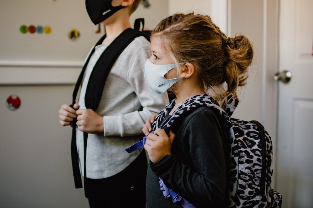 A couple of children wearing masks while standing in a home with their bags