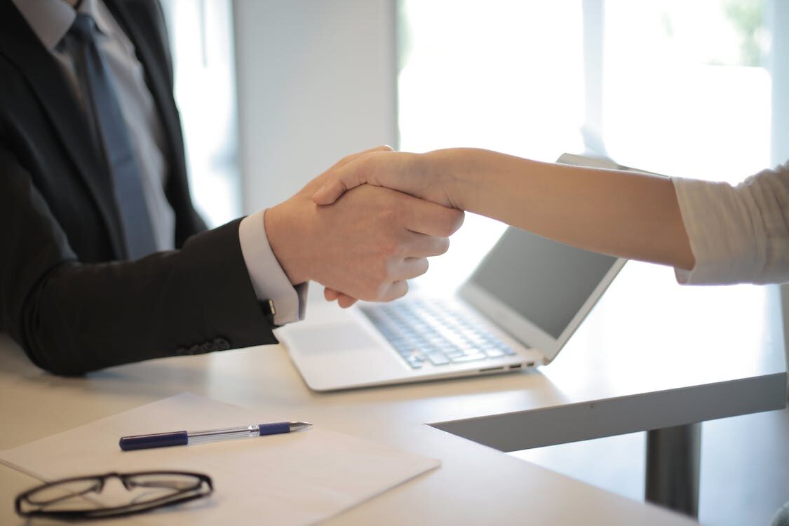 A person sitting at a desk in front of a laptop shaking hands with another person