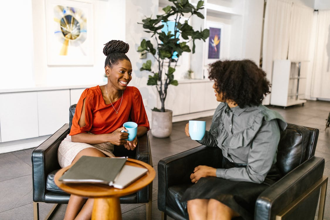 A couple of women sitting in office chairs holding coffee mugs while talking